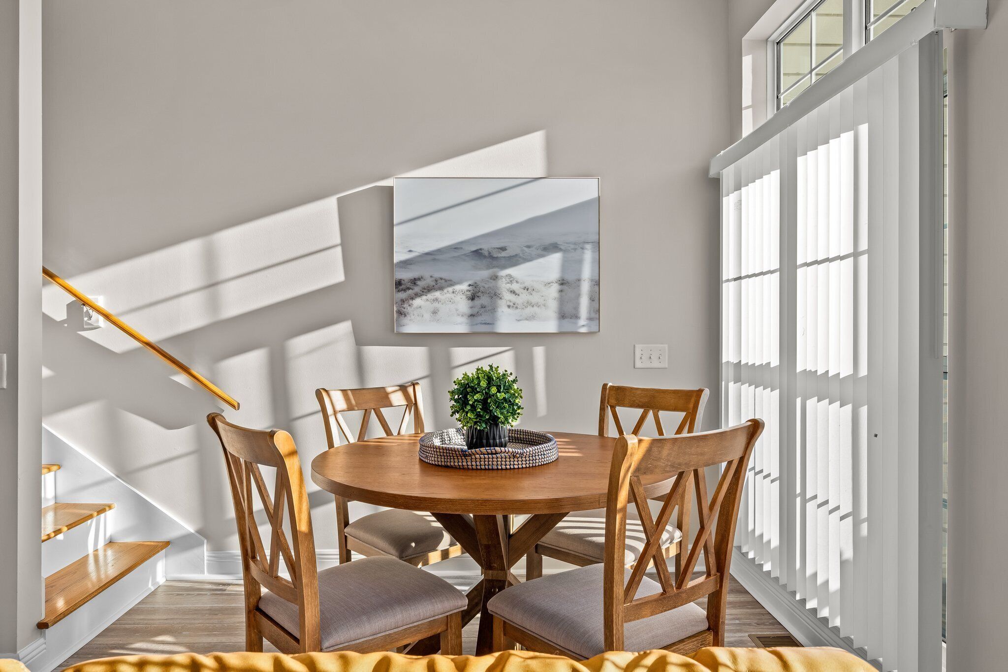 A dining area with a round wooden table, four wooden chairs, a small potted plant, and a framed picture on the wall.