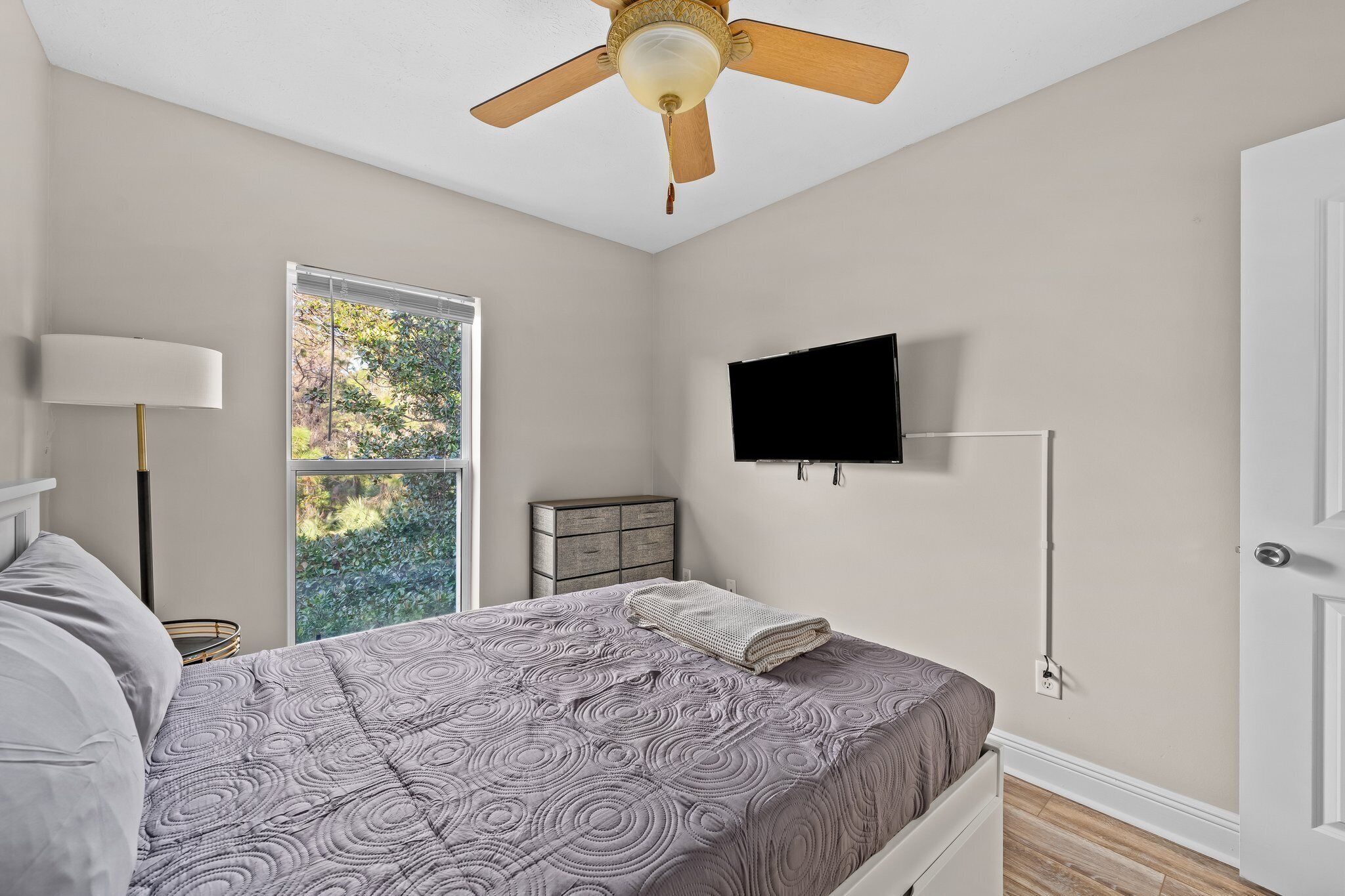 A bedroom with a bed, a ceiling fan, a TV mounted on the wall, a window with a view of greenery, and a bedside table with a lamp.