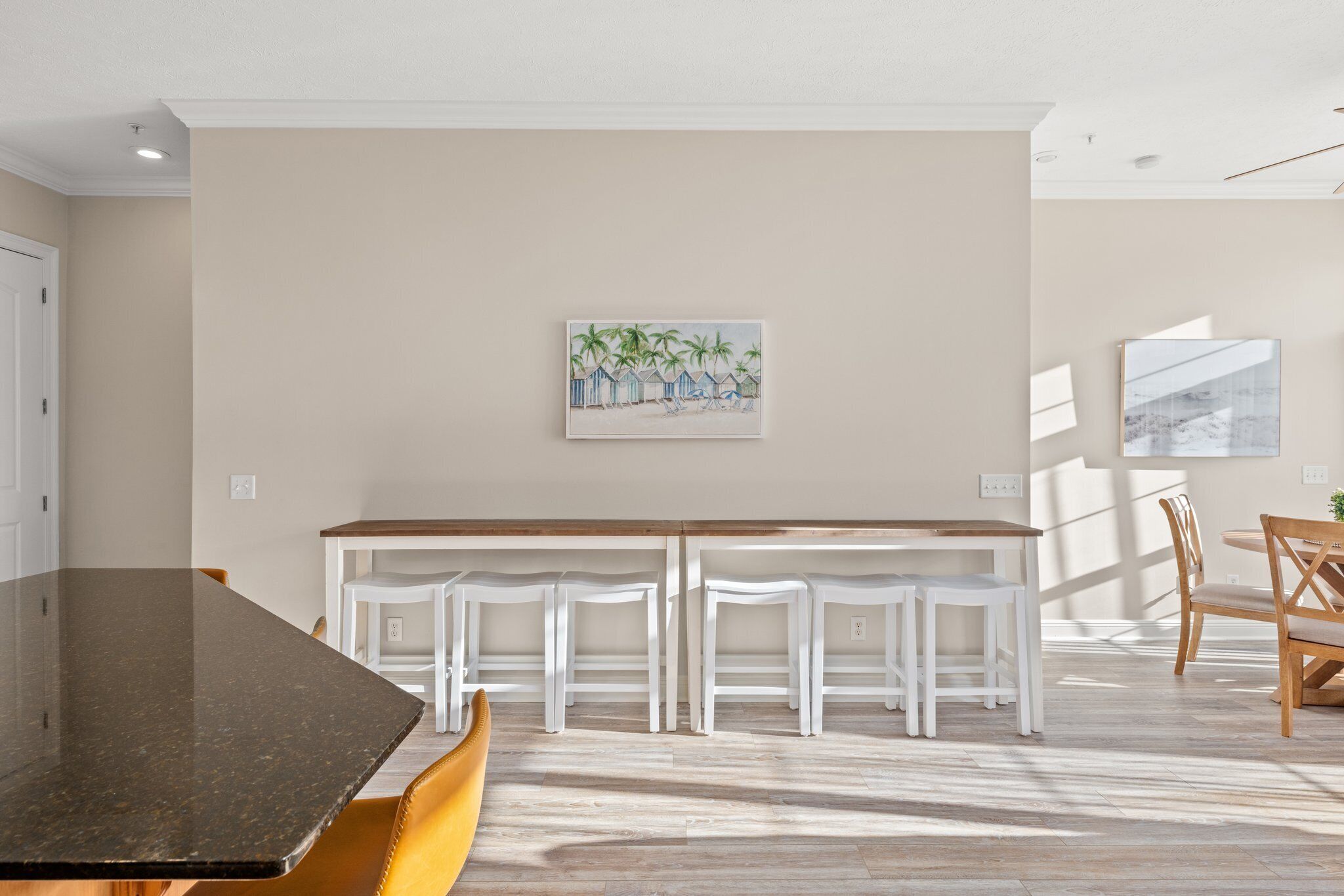 A dining area with a long table, white chairs, and a bench. There is a painting on the wall and a window providing natural light.