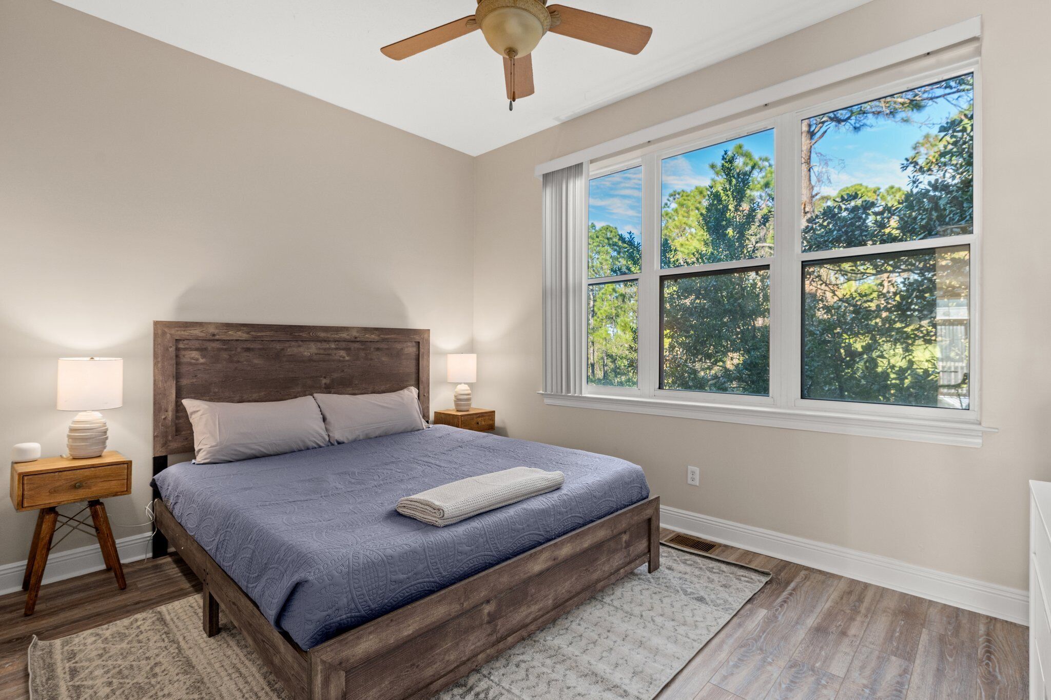 A bedroom with a wooden bed, two bedside tables with lamps, a ceiling fan, and a large window with a view of trees.