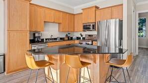 A modern kitchen with wooden cabinets, stainless steel appliances, a central island with bar stools, and a view to the outside.