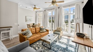 A spacious living room with a leather sofa, a coffee table, and a dining area with a table and chairs. There are ceiling fans, large windows with curtains, and a view of the city.