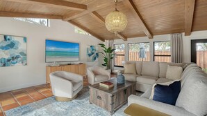 Een ruime woonkamer met een grote bank, twee fauteuils, een salontafel en een televisie. Er is een houten plafond, een groot raam en een kamerplant.