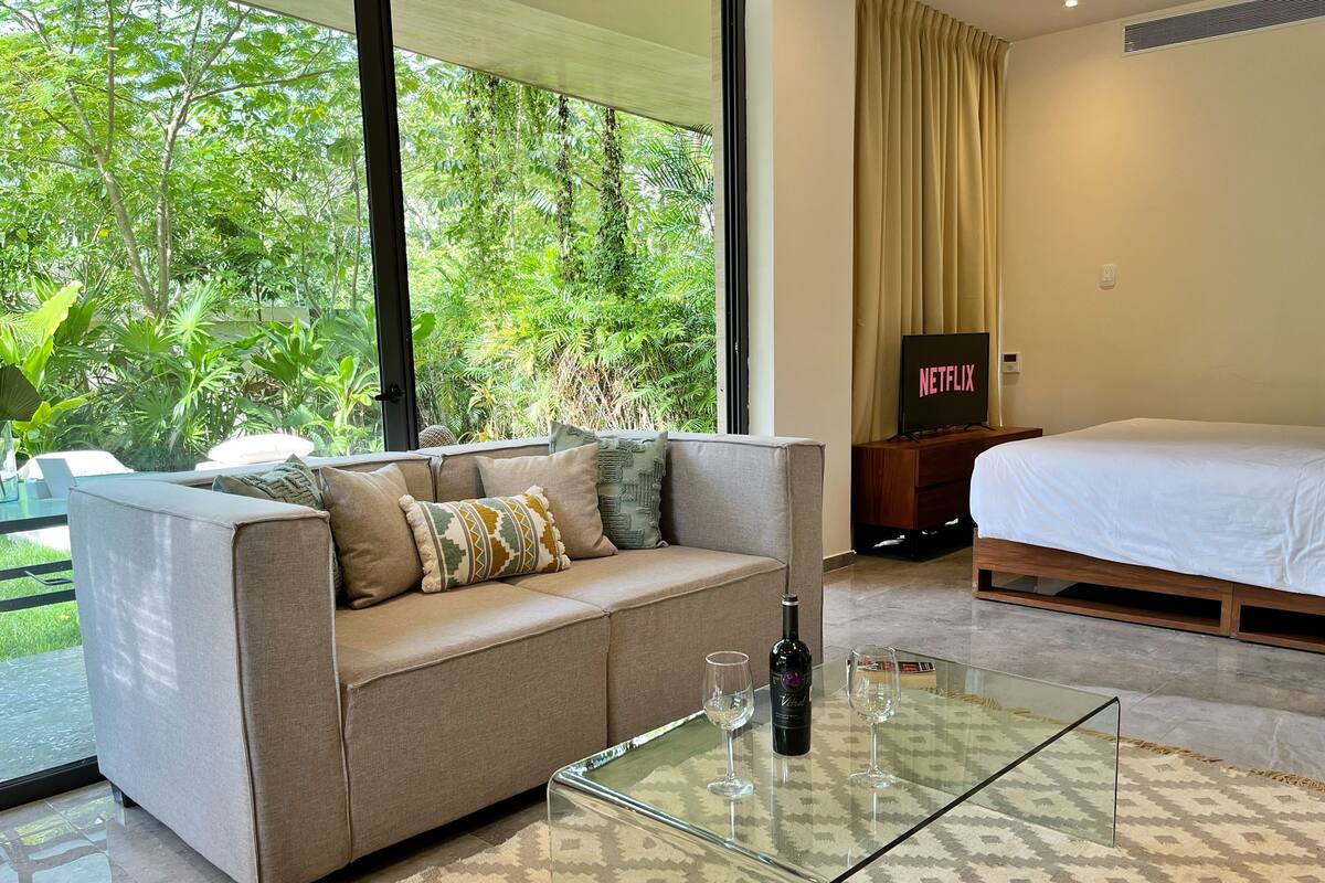 A modern hotel room with a sofa, a glass coffee table, a bed, and a TV displaying the Netflix logo.