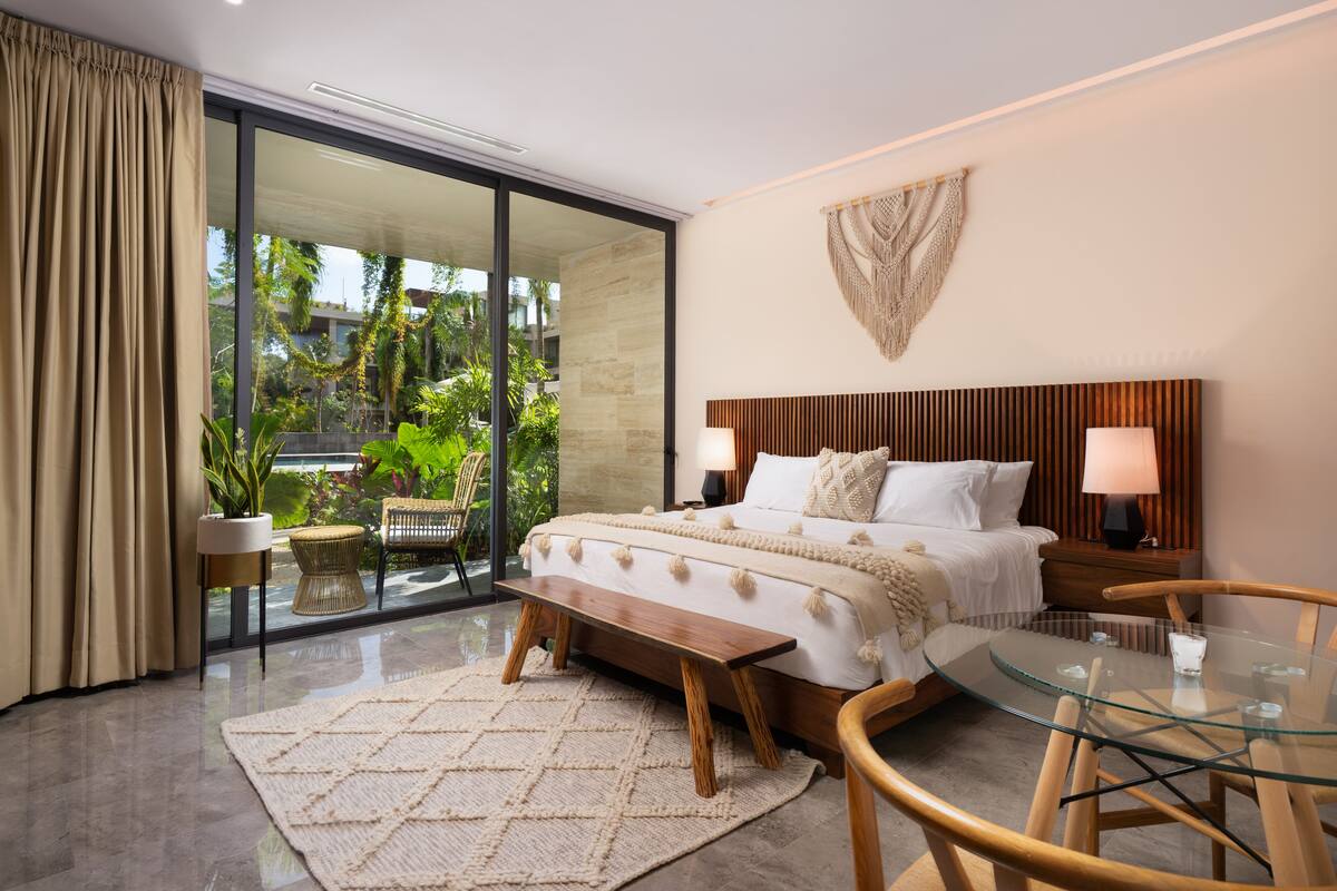 A modern bedroom with a large bed, a glass table, and a view of greenery outside.