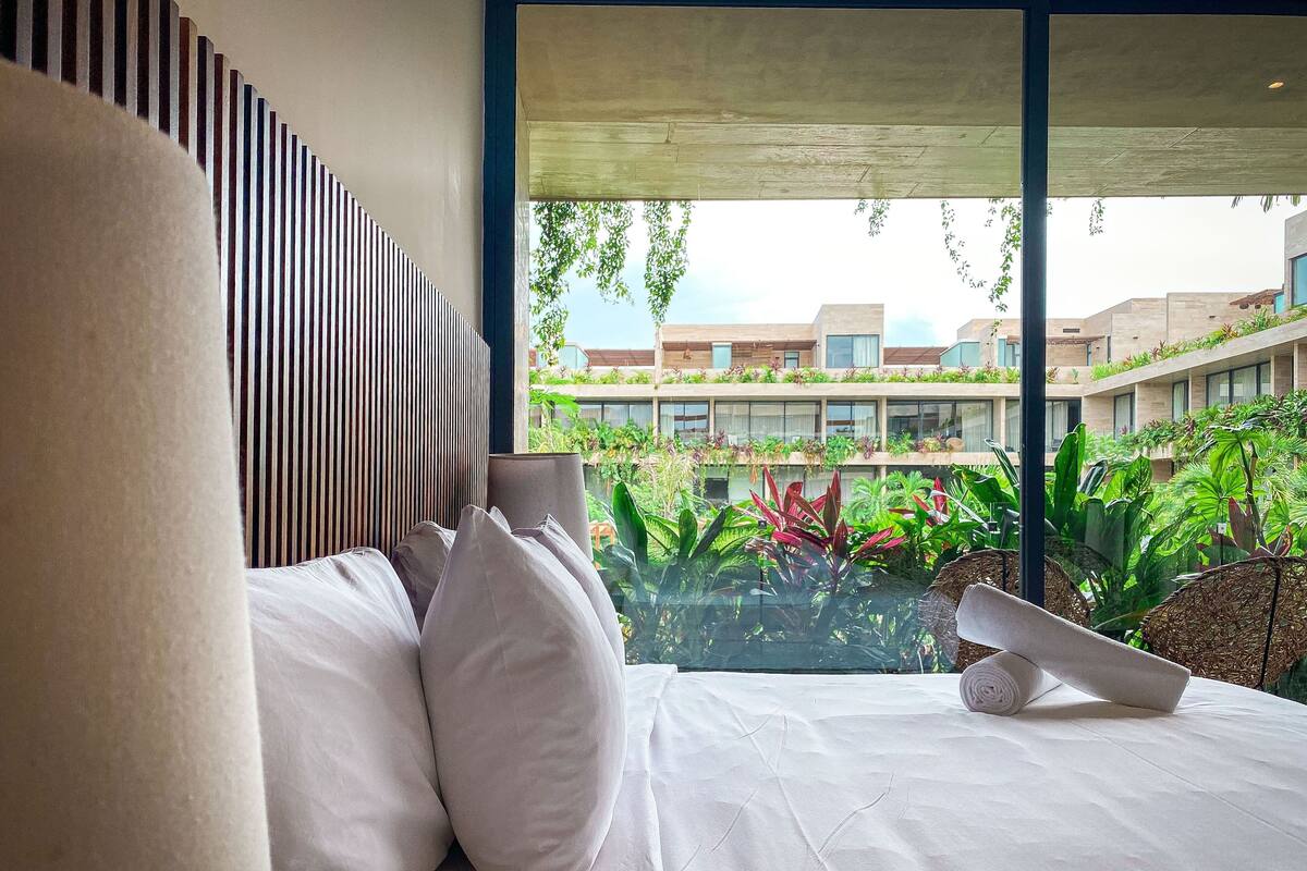 A modern hotel room with a large bed, white linens, and a view of a landscaped outdoor area with plants and buildings.