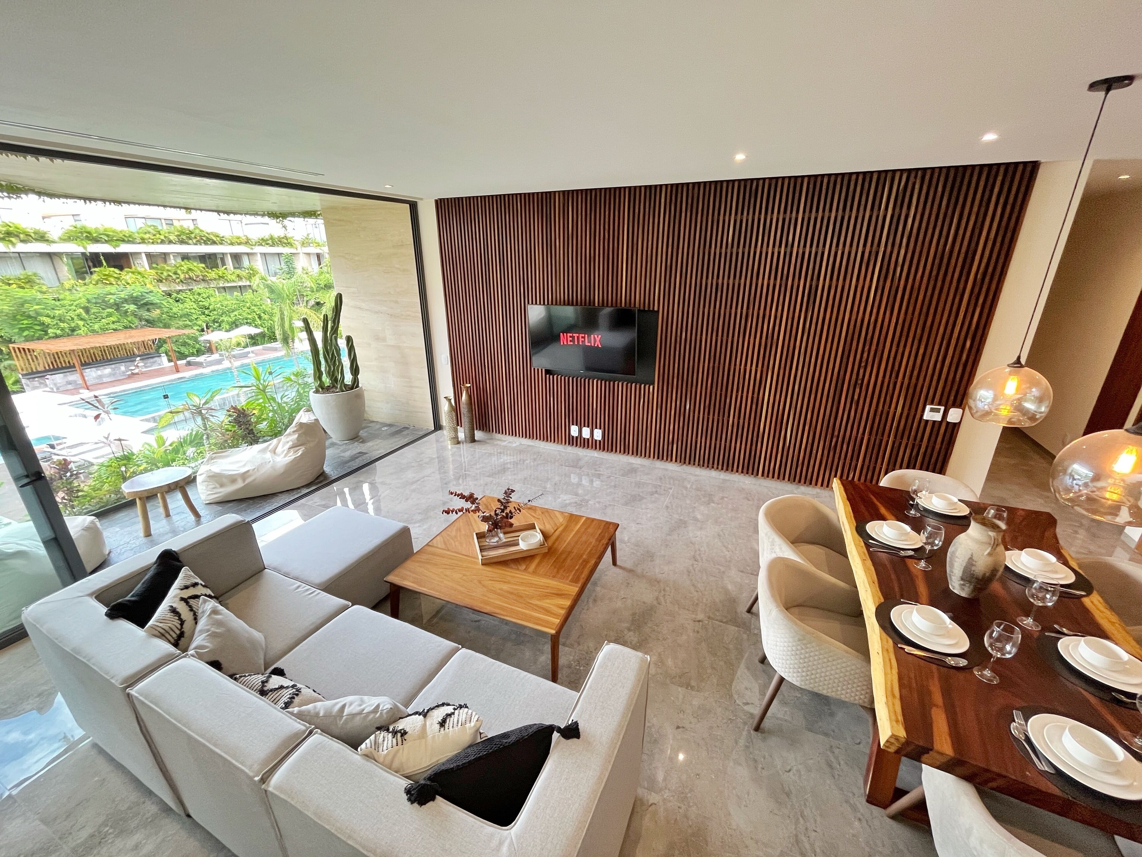 Una sala de estar moderna con un sofá, una mesa de centro y una mesa de comedor preparada para seis personas. Un televisor de pantalla plana está montado en la pared, y se ve una piscina a través de las puertas de vidrio.