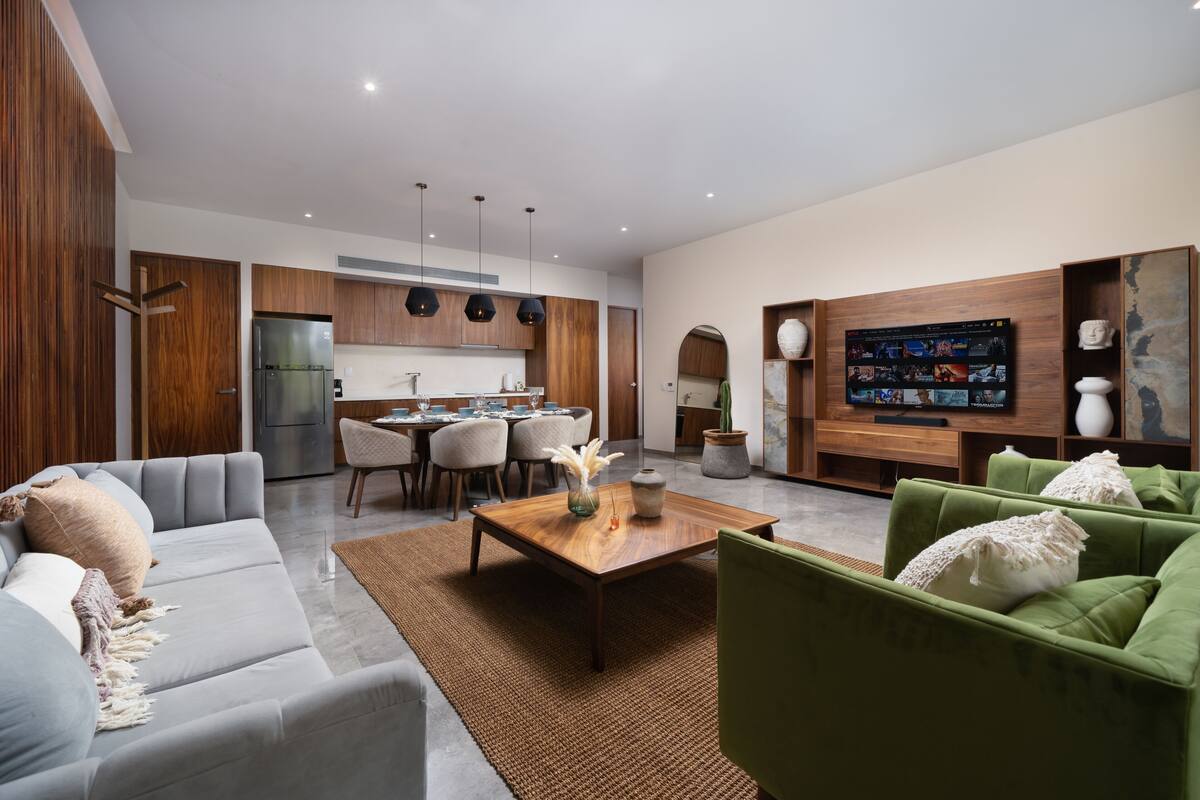 A modern living room with a grey sofa, green armchairs, a wooden coffee table, and a TV mounted on the wall.