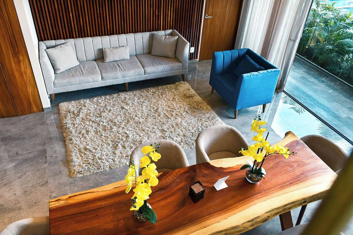 A modern living room with a sofa, a blue chair, a wooden coffee table with yellow flowers, and a view of greenery outside.