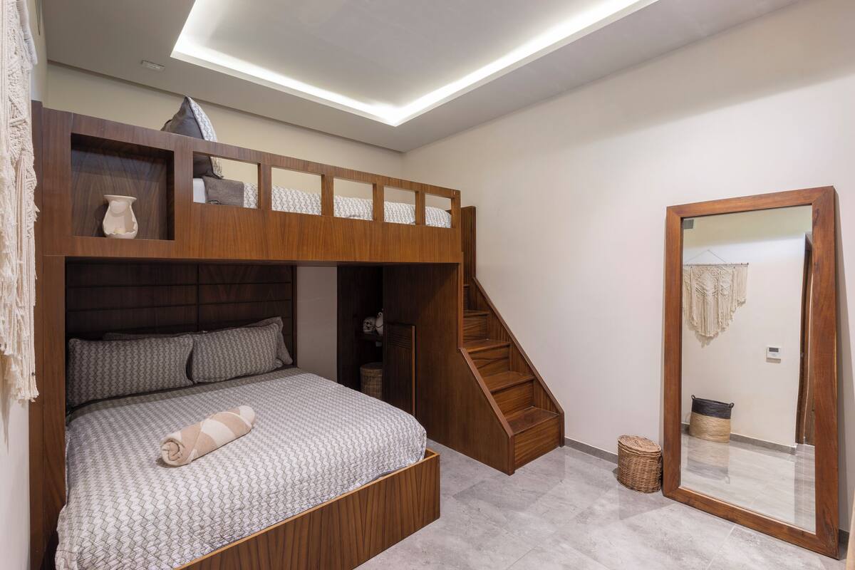 A bunk bed with a wooden headboard, a small shelf with decorative items, a mirror, and a staircase.