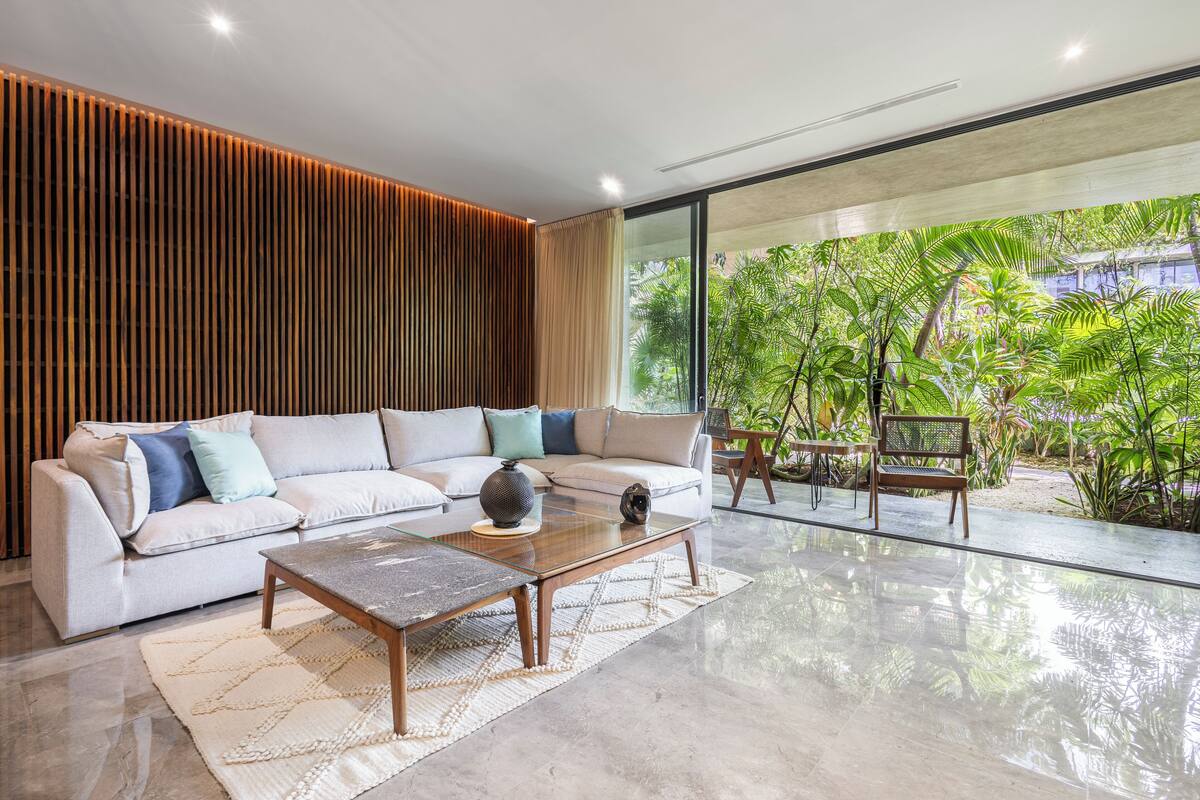 A modern living room with a sofa, coffee table, and chairs, large windows with a view of greenery.