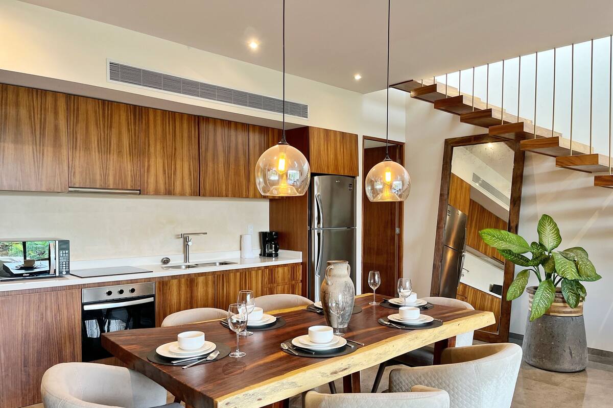 A modern kitchen with wooden cabinets, a stainless steel refrigerator, and a dining area with a wooden table and chairs.