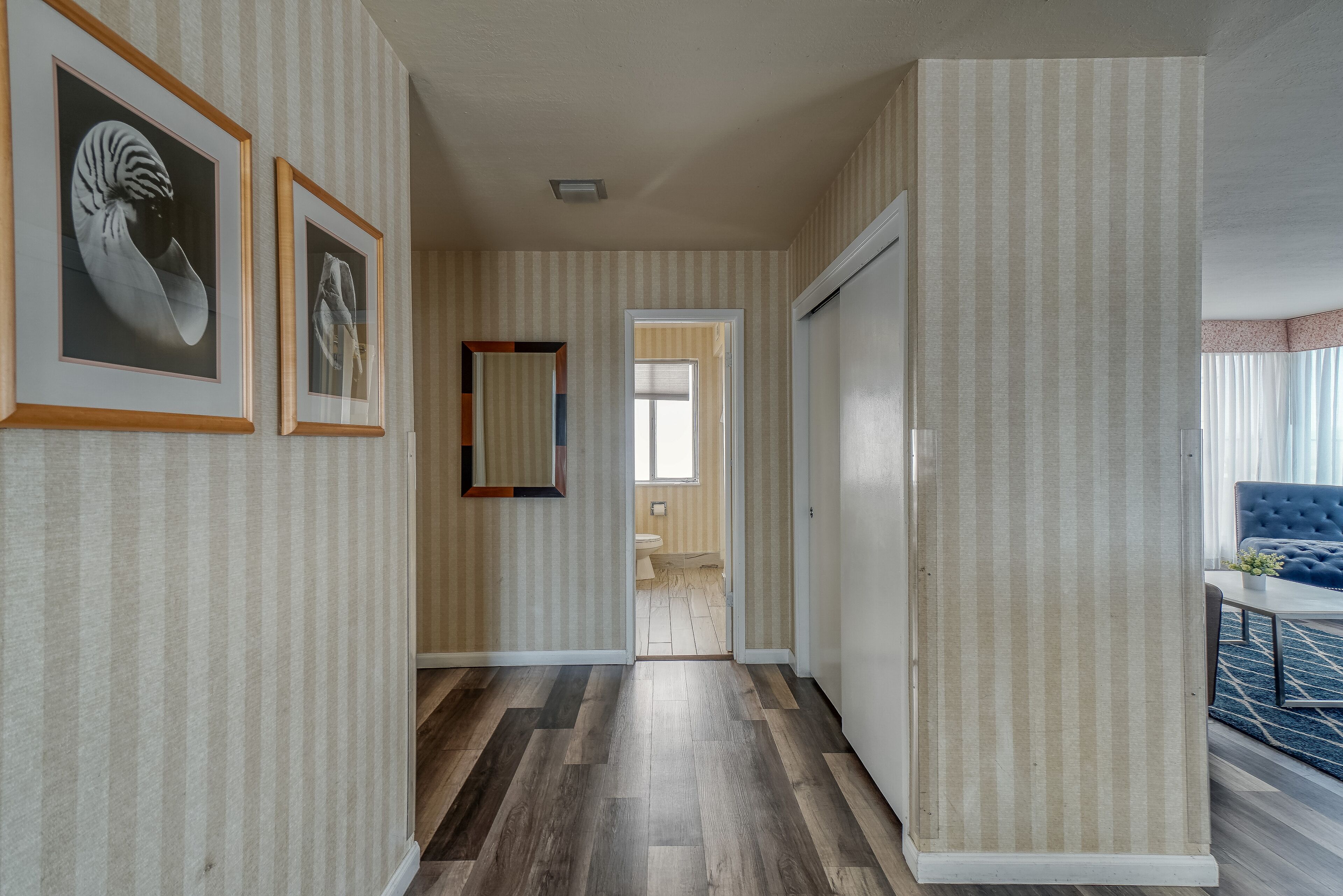 Um corredor com papel de parede listrado, quadros nas paredes, piso de madeira e vista para um banheiro.