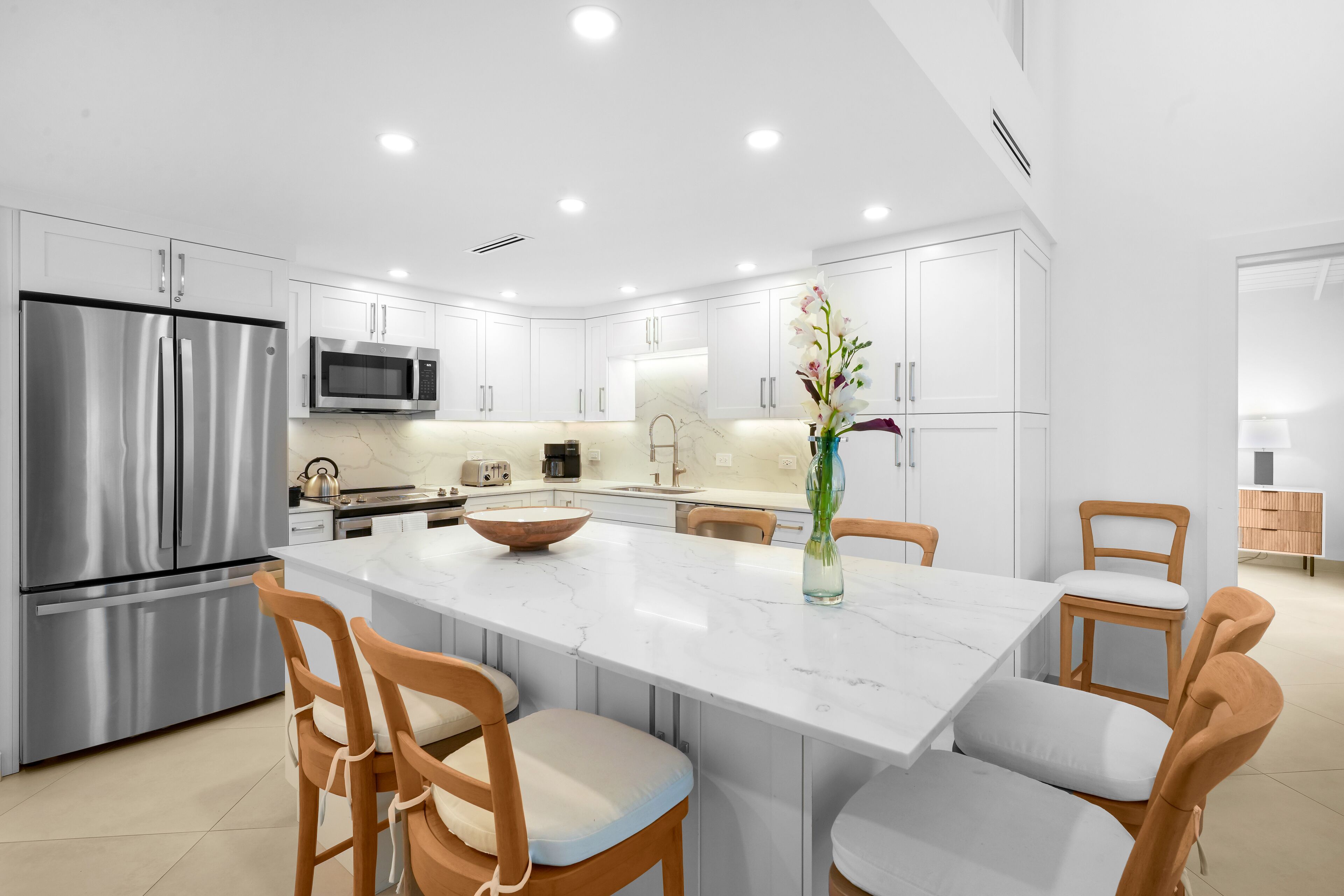 A modern kitchen with white cabinetry, stainless steel appliances, a marble island, and wooden chairs.