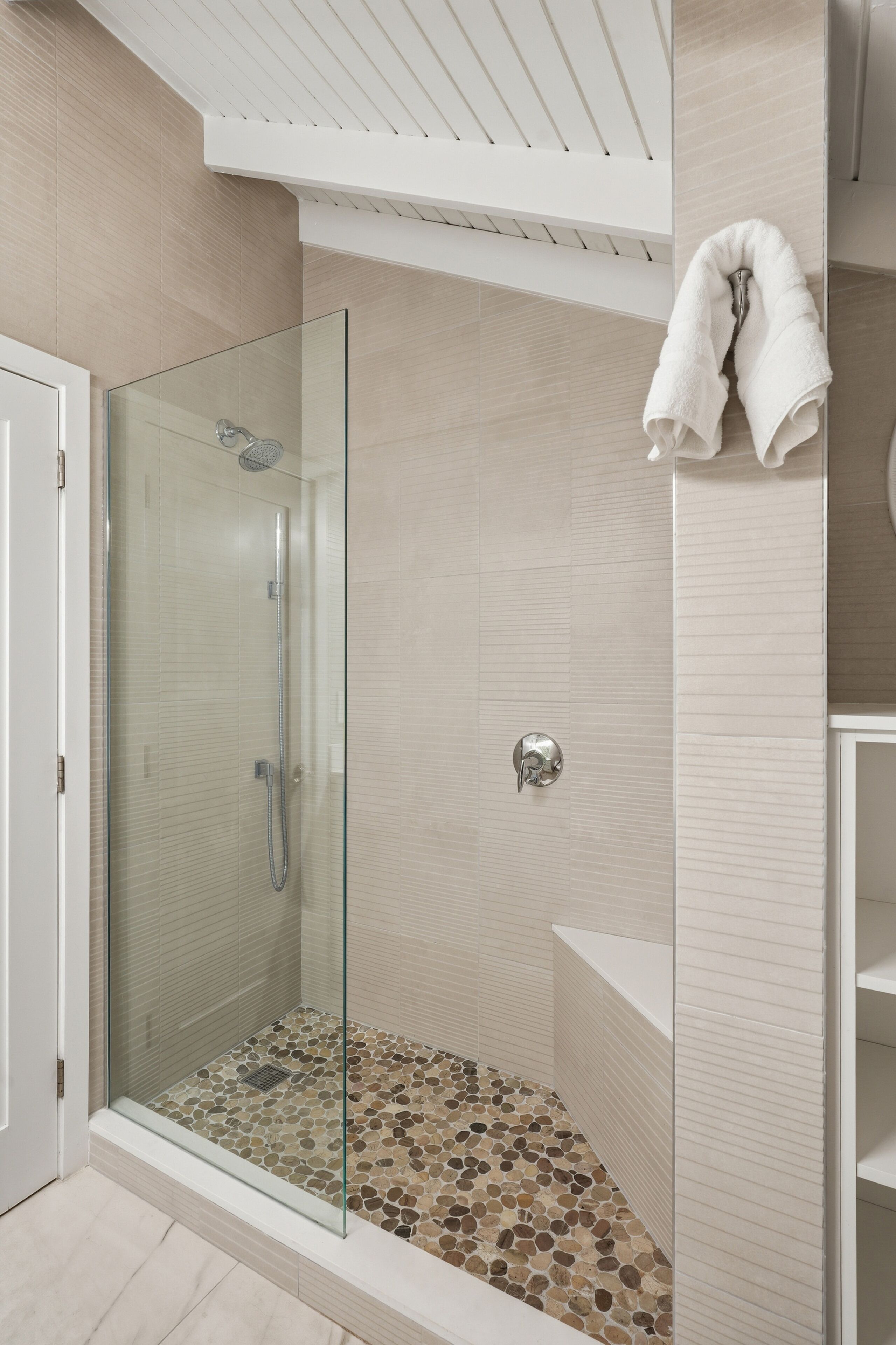 A glass-enclosed shower with a tiled floor, a white towel hanging on a hook, and a white door.