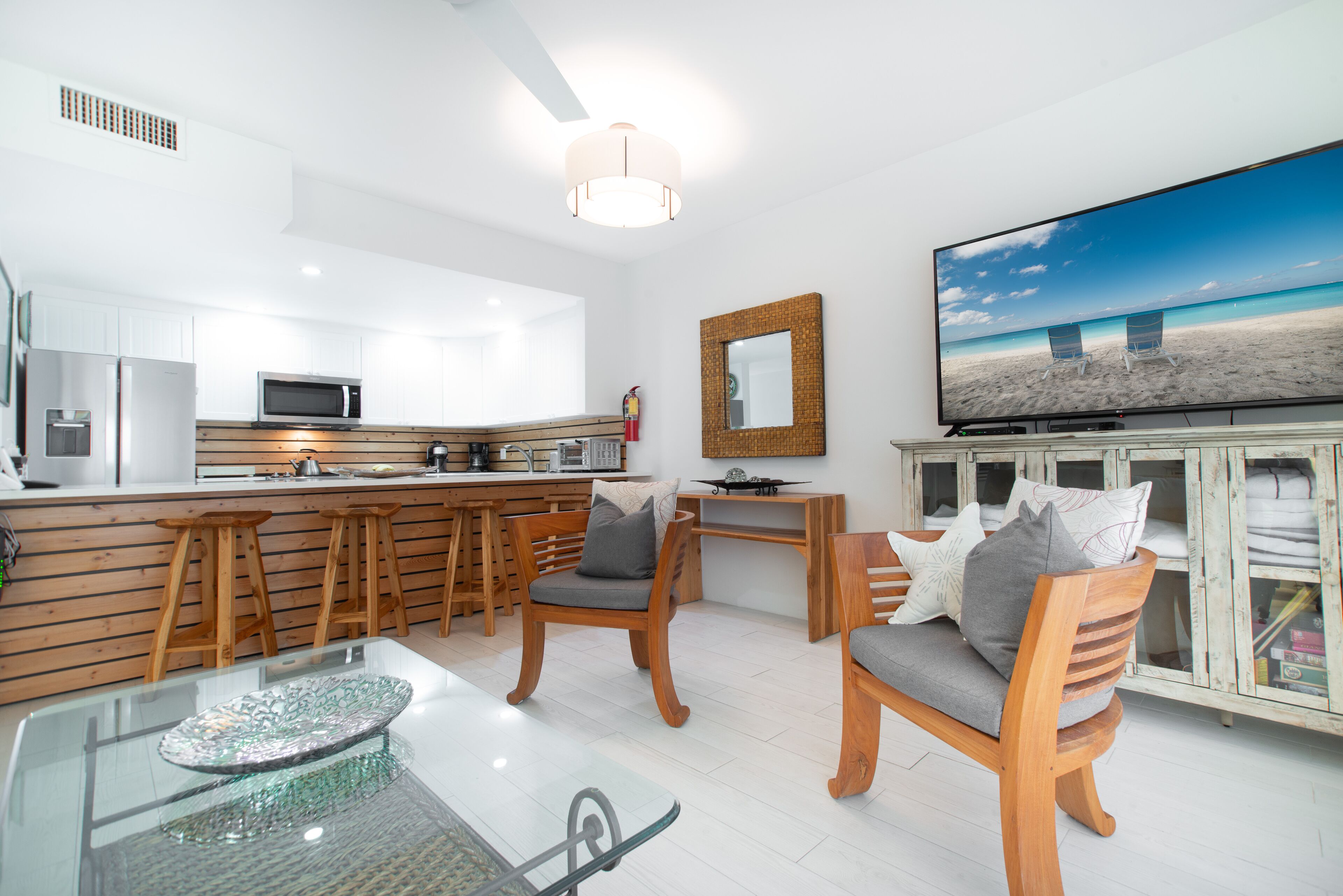 Una cocina moderna con armarios blancos, un microondas y taburetes de madera. Hay una mesa de vidrio con un plato decorativo, dos sillas de madera con cojines y un televisor de pantalla plana grande sobre un mueble.
