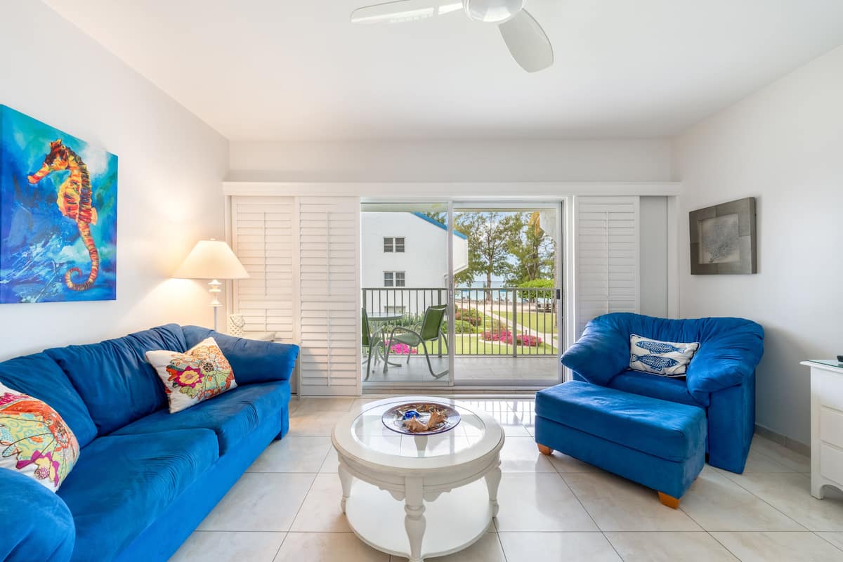 Un salon moderne avec un canapé bleu, un fauteuil assorti et une table basse blanche. Un tableau orne le mur, et on aperçoit un balcon aménagé avec du mobilier d’extérieur.