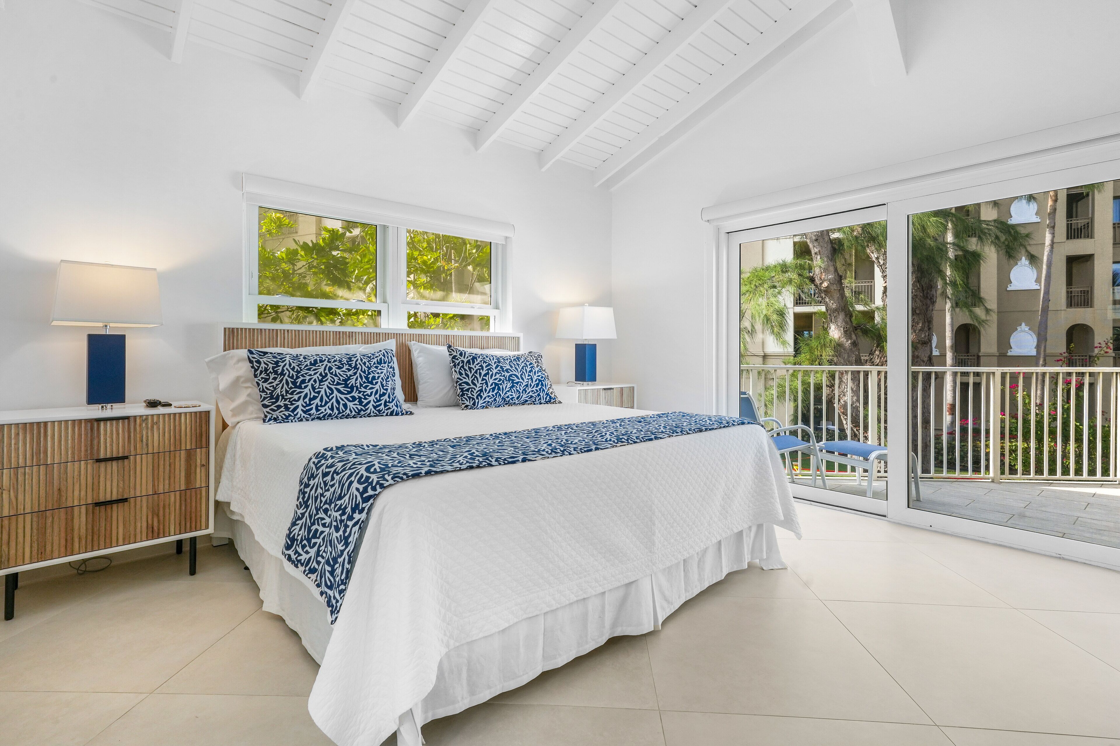 A bedroom with a large bed, two bedside tables, a window, and a balcony with a view of palm trees.