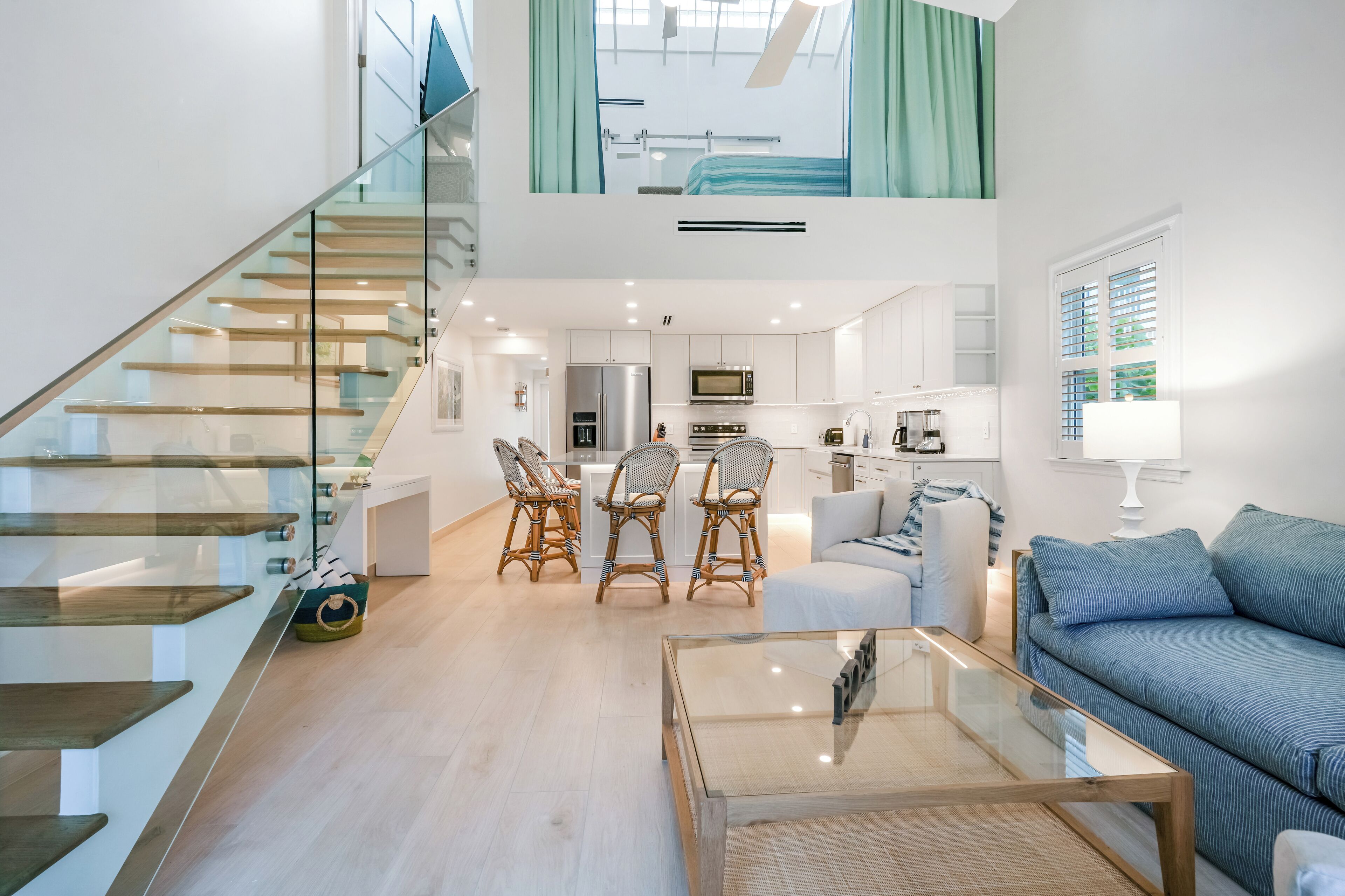 Un espacio habitable moderno con una escalera de cristal, un área de comedor con sillas de mimbre, una cocina con armarios blancos y un sofá con cojines azules.