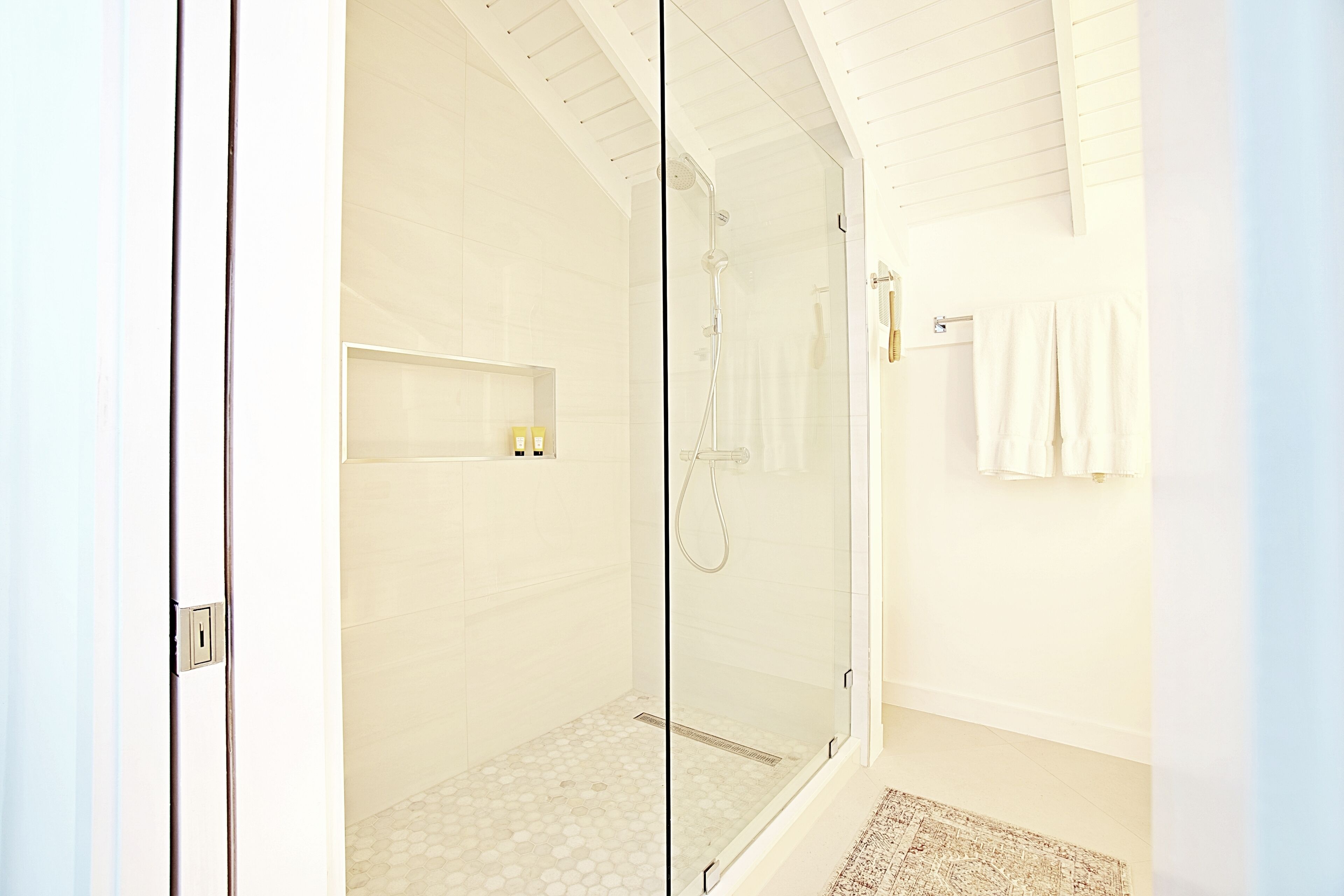 A modern bathroom with a glass-enclosed shower, white tiled walls, and a small shelf with toiletries.