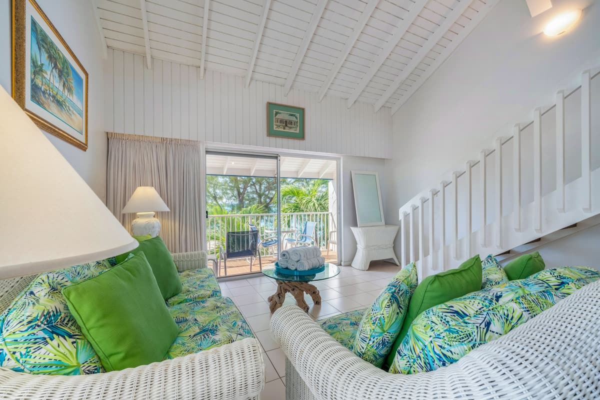 Une pièce spacieuse, meublée de mobilier en osier blanc, agrémentée de coussins verts, d’une table en verre ornée d’un élément décoratif central, et offrant une vue sur l’extérieur grâce à une porte-fenêtre coulissante.