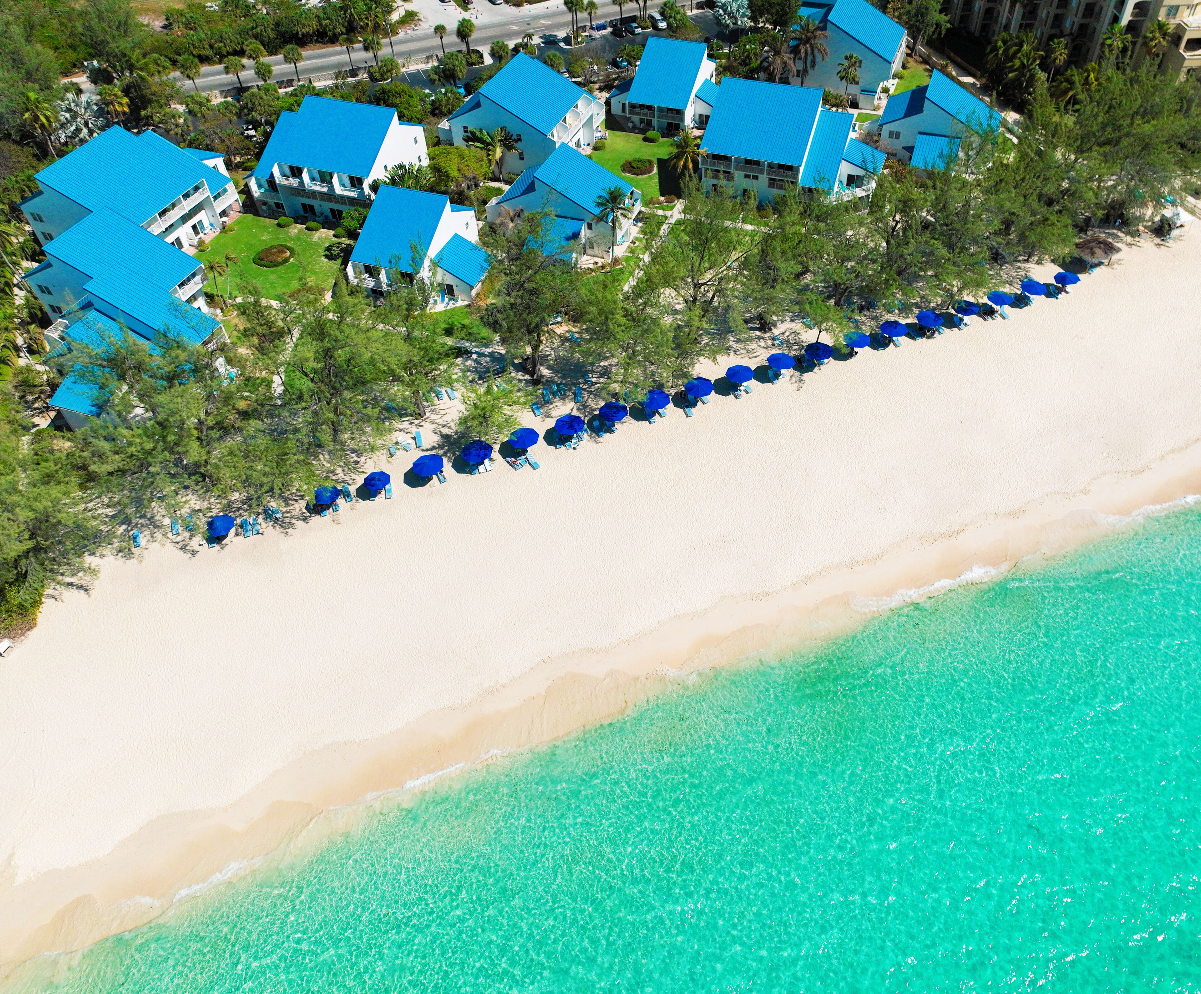 An aerial view of a beachfront property with multiple buildings, a row of beach umbrellas, and a clear turquoise sea.