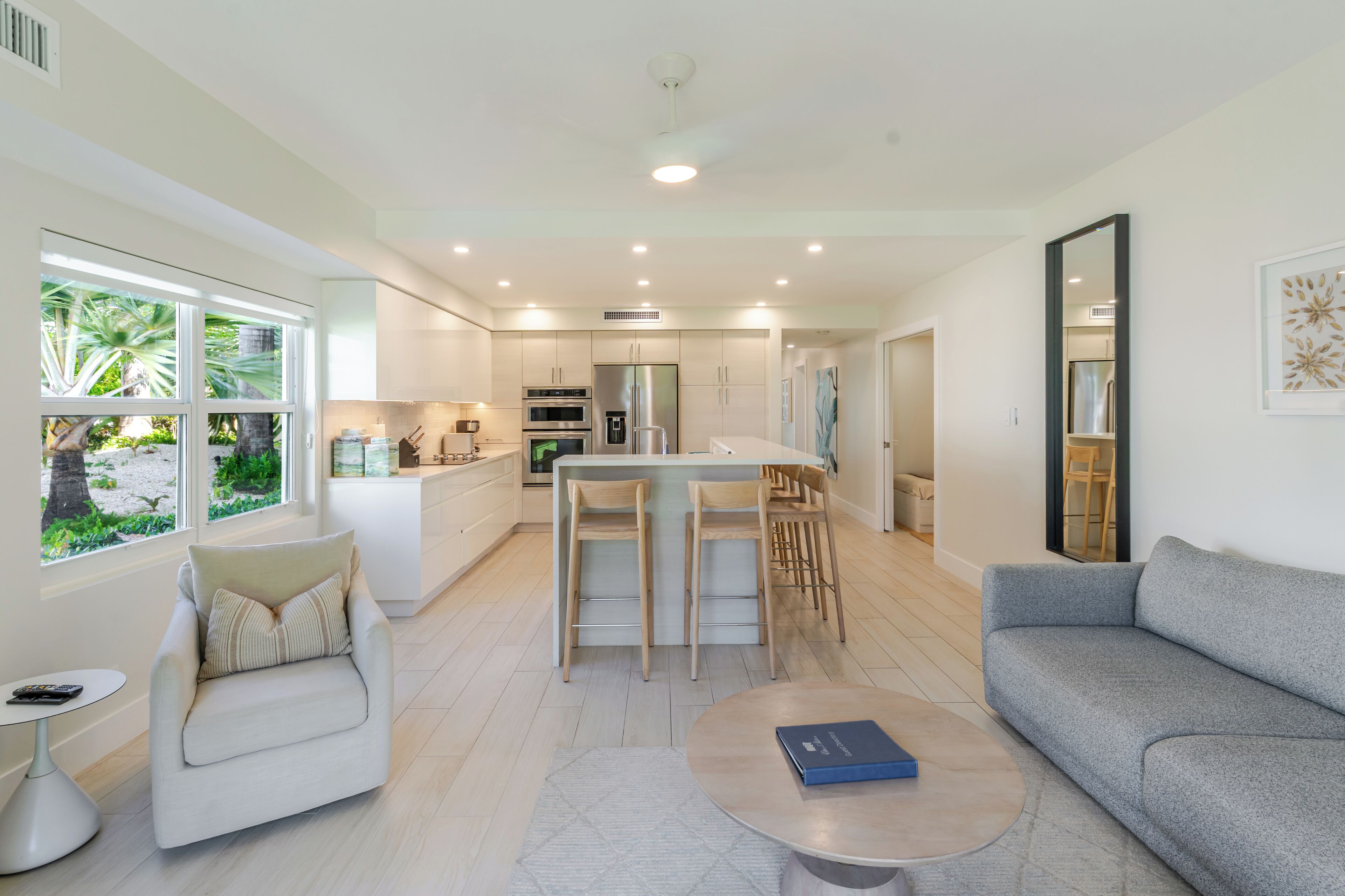Una sala de estar moderna con un sofá gris, una mesa de centro redonda y un sillón blanco. Se ve un área de cocina con una isla central y taburetes de bar al fondo.