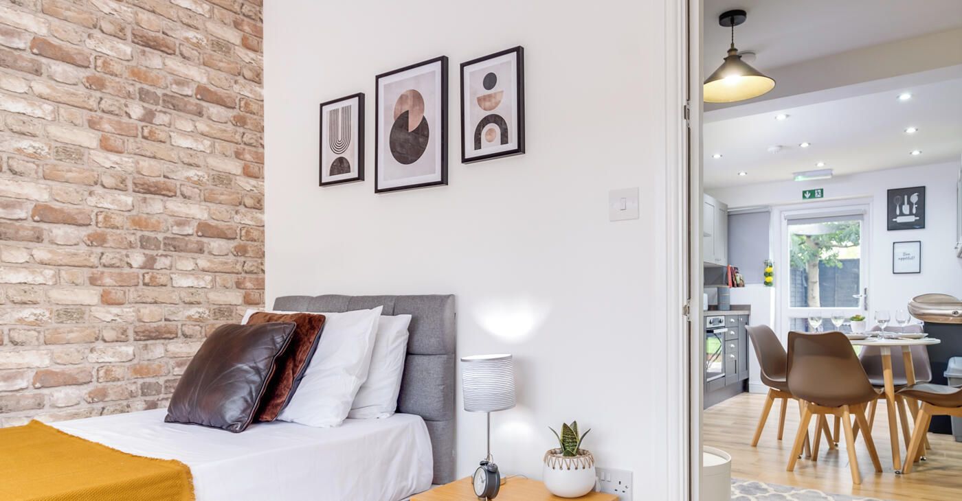A modern bedroom with a brick wall, a bed with multiple pillows, a bedside table with a lamp and a plant, and a view into a dining area with a table and chairs.