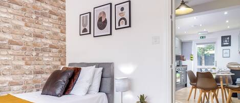 A modern bedroom with a brick wall, a bed with multiple pillows, a bedside table with a lamp and a plant, and a view into a dining area with a table and chairs.