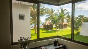 A room with a large window offering a view of a lawn with palm trees and a thatched-roof structure. - Beach house w/ private pool & sandy beach for 8 (San Miguel de Cozumel)