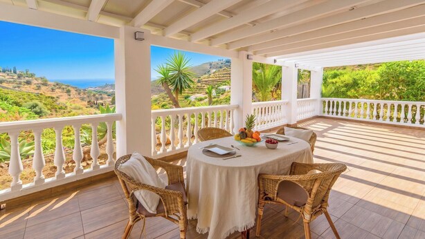 A covered outdoor seating area with wicker furniture, a round table with a white tablecloth, and a view of the landscape.