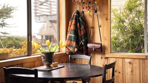 A cozy room with a wooden interior, a round table with chairs, a window with a view of greenery, and a hanging light fixture. - Sundog Cottage - Clean & Cozy - EV - Walk to Food & Beach - Fenced Yard for Dog (Dillon Beach)