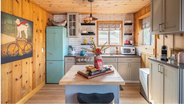 A kitchen with wooden walls, a blue refrigerator, a wooden island with a vase and coffee maker, and a window with a view. - Sundog Cottage - Clean & Cozy - EV - Walk to Food & Beach - Fenced Yard for Dog (Dillon Beach)