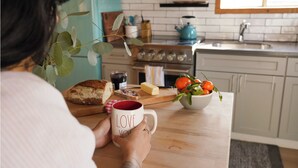 A person holding a mug with "LOVE YOU" written on it, sitting at a wooden kitchen table with bread, a bowl of fruit, and a blue kettle in the background. - Sundog Cottage - Clean & Cozy - EV - Walk to Food & Beach - Fenced Yard for Dog (Dillon Beach)