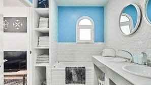Bagno con piastrelle bianche e blu, una vasca, due lavabi e un televisore nell'angolo.