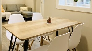 A dining area with a wooden table and white chairs, a sofa with a 'welcome' pillow, a painting of a beach scene, and a window with a plant. - Charming 2 Bedroom Apt. Close to Valparaiso University Short-Term Stays (Valparaiso)