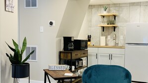 A modern kitchen with white cabinets, a microwave, and a coffee maker on a wooden table. There is a blue chair and a potted plant in the foreground. - Charming 2 Bedroom Apt. Close to Valparaiso University Short-Term Stays (Valparaiso)