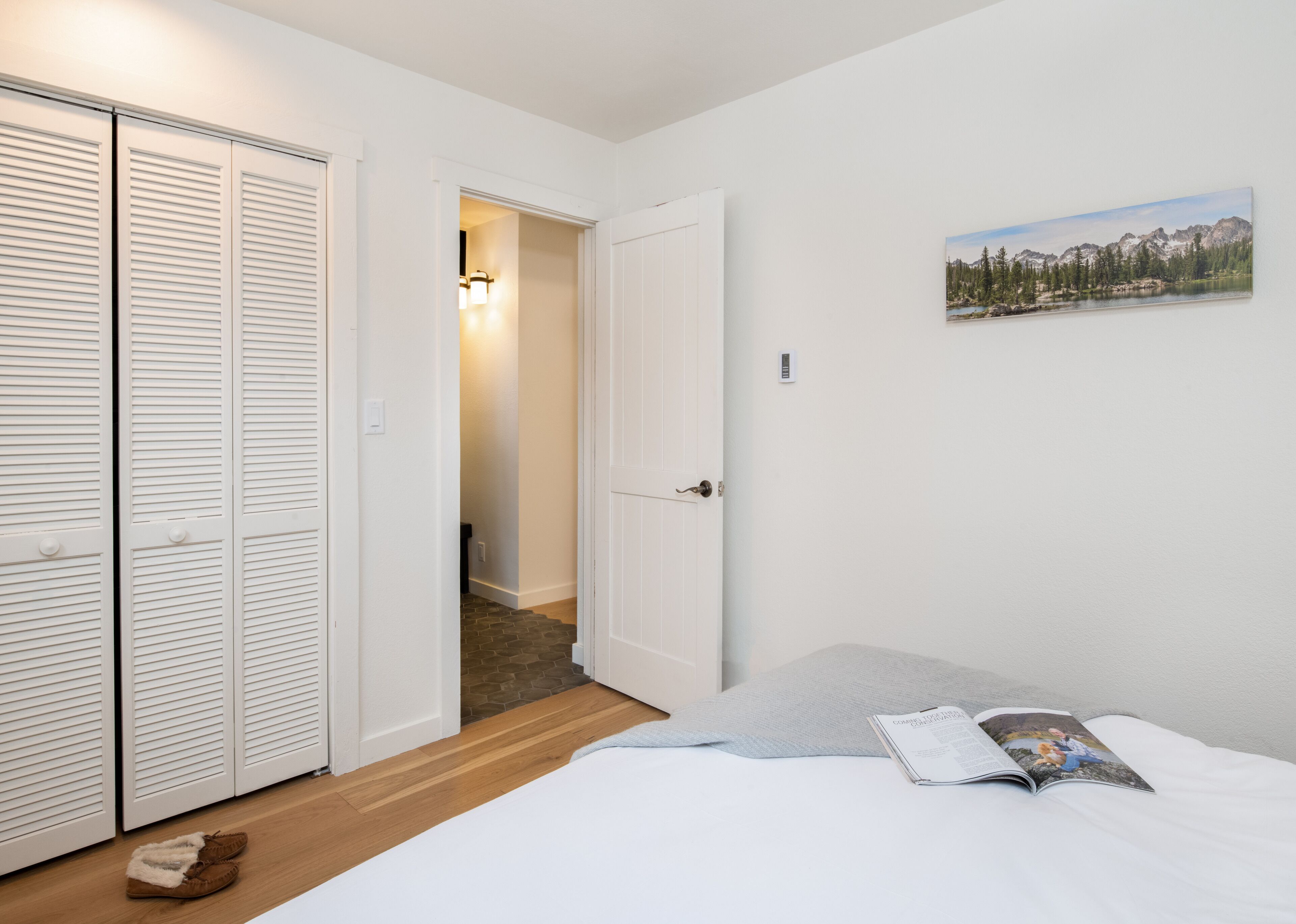 A bedroom with a bed, a magazine, a pair of slippers, and a painting of a mountain landscape on the wall.