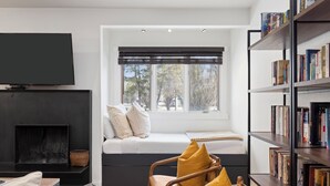 A modern living room with a sofa by a large window, a bookshelf, and a flat-screen TV mounted on the wall. - Outpost: Cedars 1413 - Aspens Location! (Wilson)
