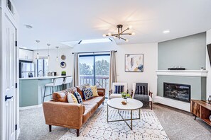 A modern living room with a sofa, chairs, a fireplace, and a kitchen in the background. - Modern DTC 1BR Condo with Pool, Fireplace, Light Rail (Greenwood Village)