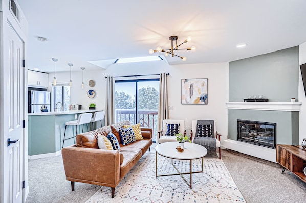 A modern living room with a sofa, chairs, a fireplace, and a kitchen in the background.