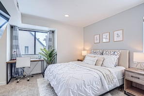 A bedroom with a bed, desk, and chair, a window with curtains, and wall-mounted artwork.