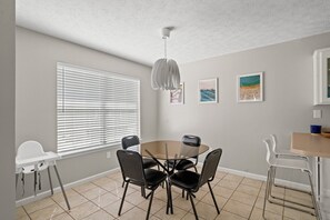 Een eetgedeelte met een ronde tafel, vier stoelen, een hoge plafond en een raam met jaloezieën.