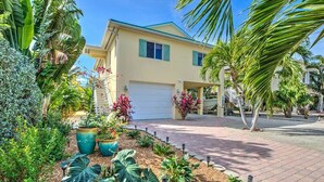 Una casa de dos pisos con garaje, rodeada de plantas tropicales y un camino de entrada pavimentado.