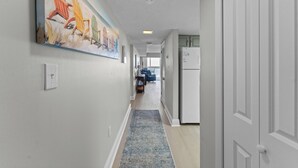 A hallway with a painting of chairs, a rug, and a refrigerator. - ☀️Edgewater West 34. stocked poolside storage room.  New paint and floors!!⛱️ (Gulf Shores)