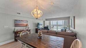 A dining area with a wooden table and chairs, a chandelier, a sofa, and a television. - ☀️Edgewater West 34. stocked poolside storage room.  New paint and floors!!⛱️ (Gulf Shores)