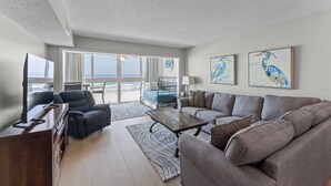 A living room with a sofa, armchairs, a coffee table, and a TV. There are two paintings on the wall and a view of the ocean through large windows. - ☀️Edgewater West 34. stocked poolside storage room.  New paint and floors!!⛱️ (Gulf Shores)