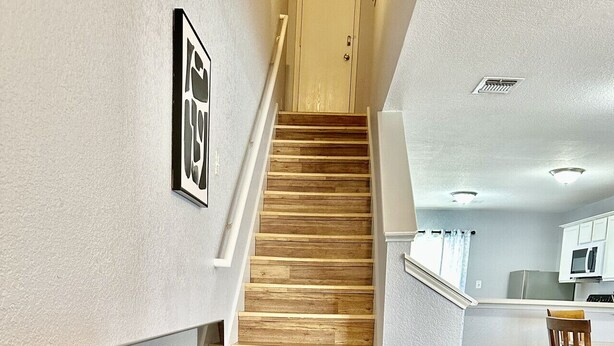Un interior moderno con una escalera de madera que lleva a un segundo piso, una zona de comedor con mesa y sillas, y una cocina con armarios y electrodomésticos.
