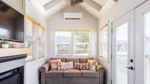 A cozy living room with a grey sofa, two windows with blinds, a flat-screen TV, a built-in oven, and a ceiling fan. - Zion-Day-Trip Ready • Tiny Home + Mountain Views (Apple Valley)