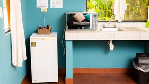 Une cuisine compacte équipée d’un four à micro-ondes, d’un évier et d’un petit réfrigérateur. Deux tableaux sont accrochés au mur, et une fenêtre offre une vue sur la verdure.